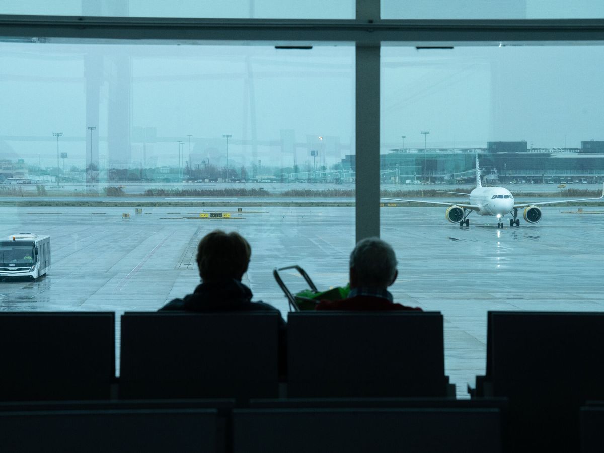 Foto: Dos personas esperan sentadas frente a un avión aparcado en la pista del aeropuerto de El Prat. (Europa Press/David Zorrakino)