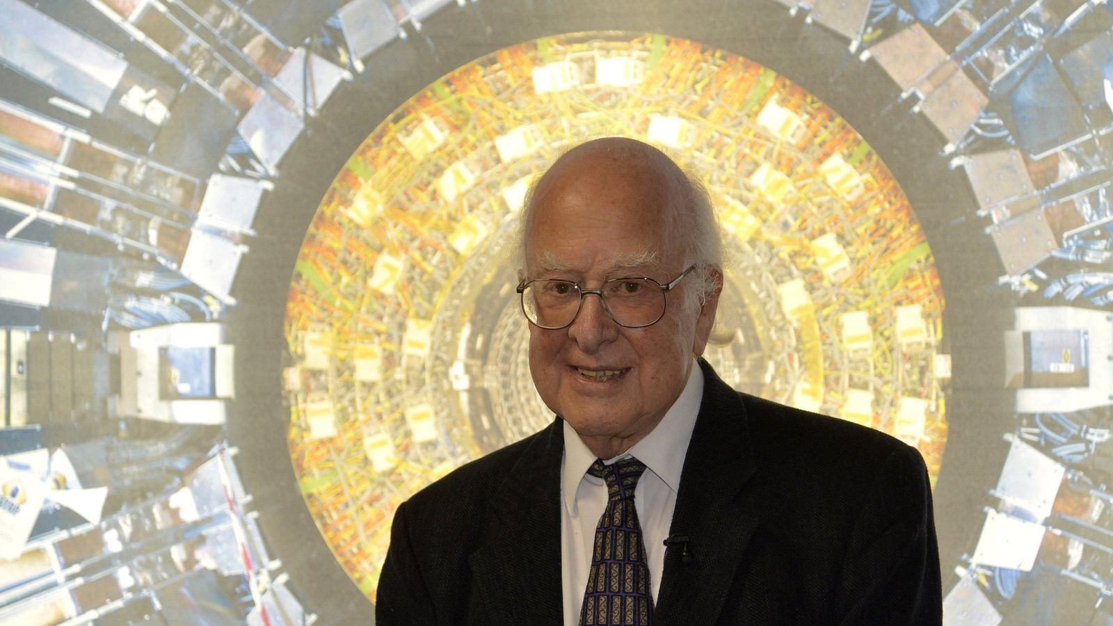 El científico Peter Higgs posando junto a una fotografía del detector Atlas del LHC, uno de los instrumentos que confirmaron su teoría del boson de Higgs.