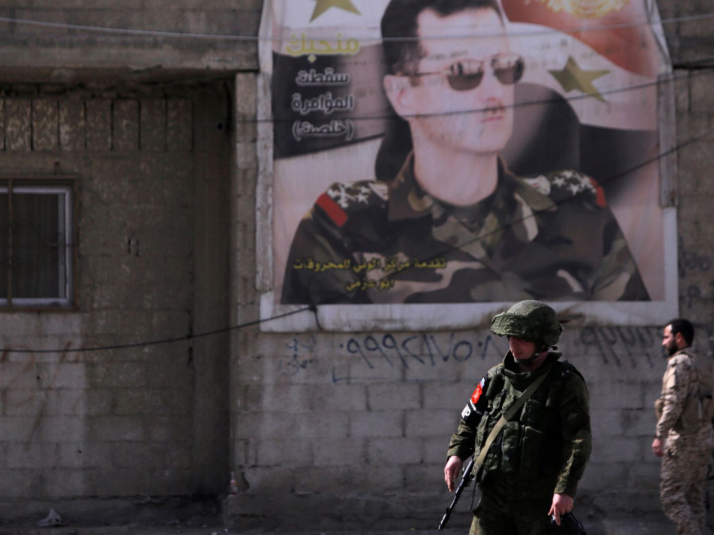 Un soldado ruso y otro sirio, frente a un retrato de Bashar al Asad en el campo de Wafideen, en Damasco, el 1 de marzo de 2018. (Reuters)