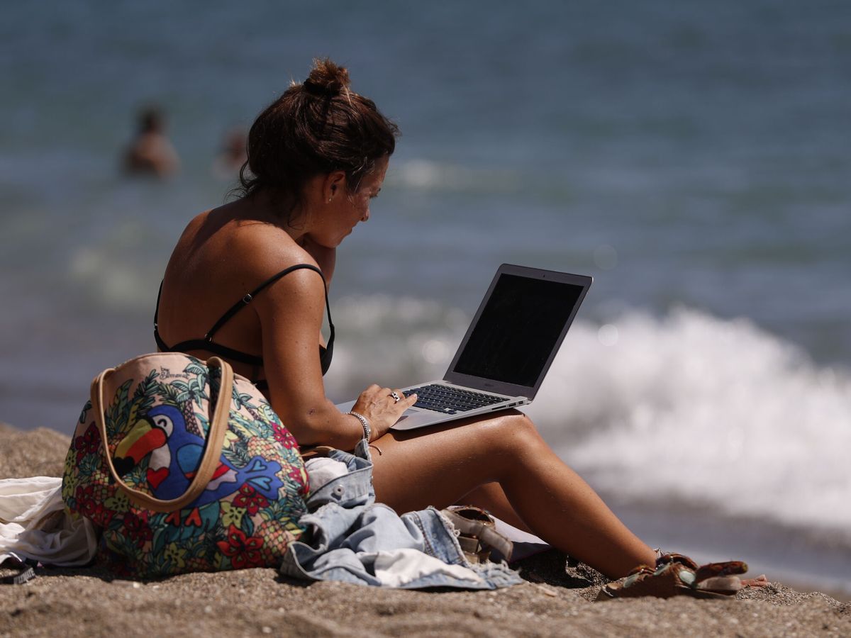Foto: Una mujer toma el sol mientras mira su ordenador portátil en la playa de la Malagueta, en Málaga, hace unas semanas. (EFE/Jorge Zapata)