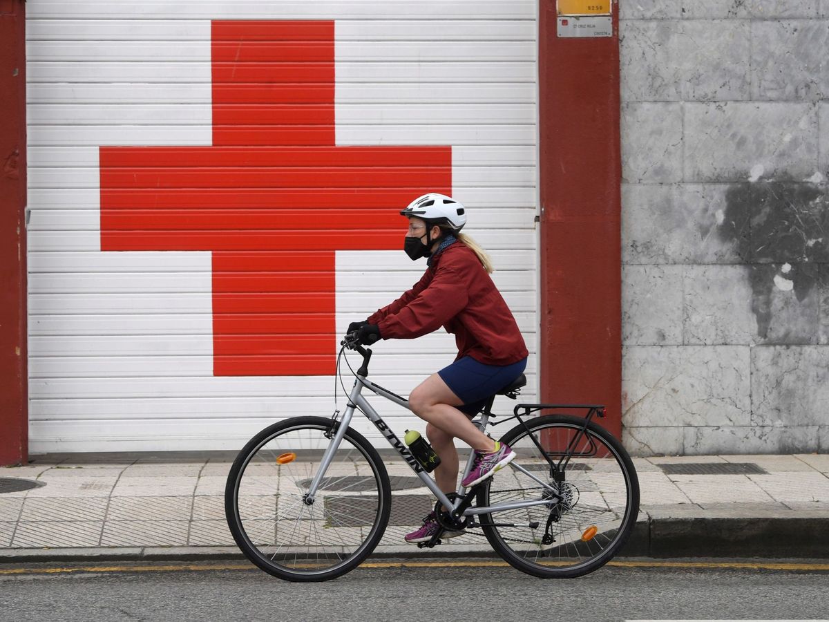 Foto: Una mujer pasea en bicicleta con mascarilla ante la puerta de entrada de ambulancias del Hospital de La Cruz Roja en Oviedo. (EFE)