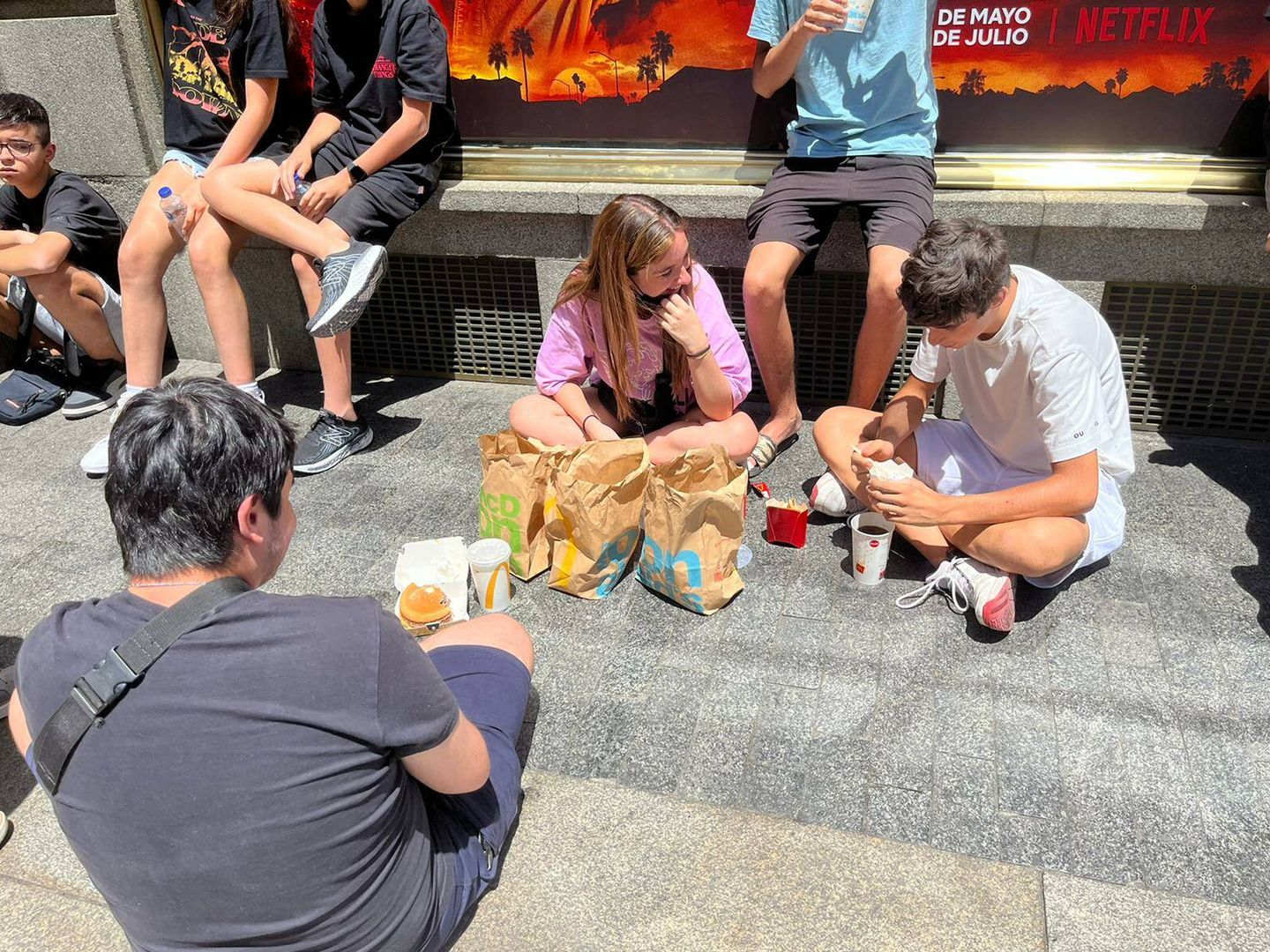 Los adolescentes comiendo en el suelo bajo el sol mientras hacen cola. (A.F.)