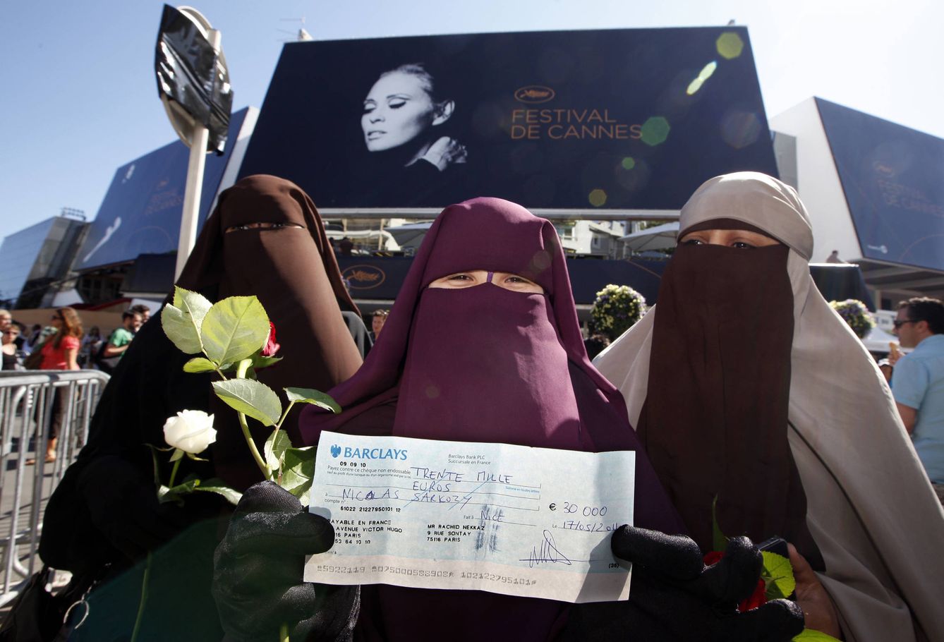 Tres mujeres con niqab posan ante una cartel del festival de Cannes con un cheque que simboliza una posible multa por llevar velo integral. (Reuters)
