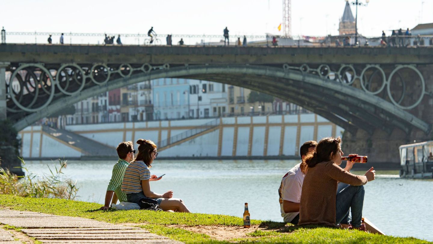 Varias personas se sientan junto a la dársena del Guadalquivir, con el puente de Triana al fondo. (EFE)