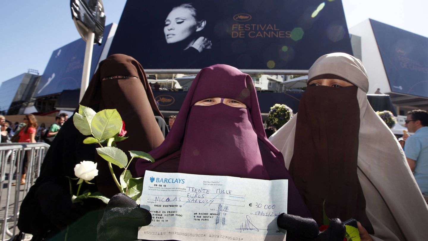 Tres mujeres con niqab posan ante una cartel del festival de Cannes con un cheque que simboliza una posible multa por llevar velo integral. (Reuters)