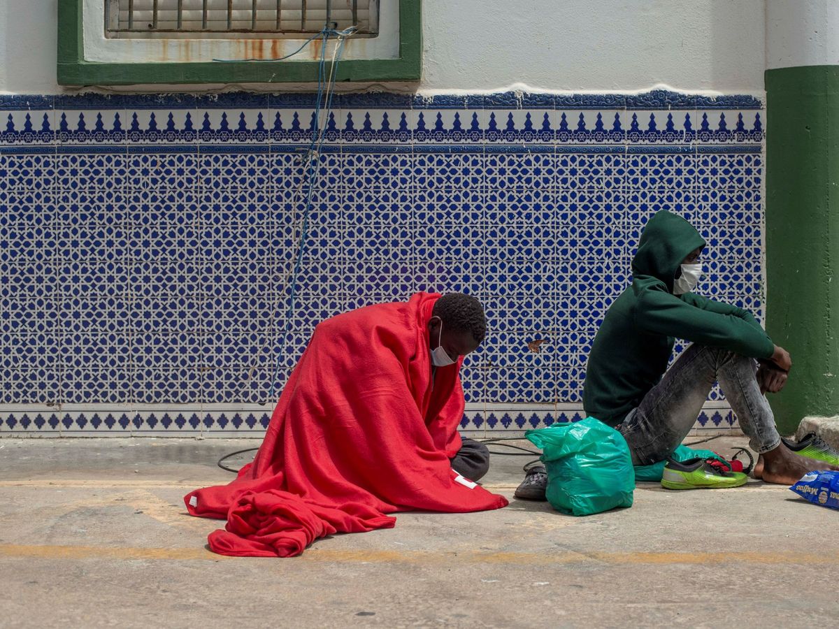 Foto: Dos migrantes que buscan asilo esperan a ser ayudados a las puertas del polígono industrial cercano a la playa del Tarajal (Ceuta). (EFE)