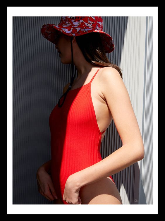 Traje de baño rojo de canalé con escote halter especialmente favorecedor para remarcar los hombros, de BERSHKA. La modelo lo lleva con un divertido sombrero estampado de BIMBA & LOLA, perfecto para las horas de sol más intensas.