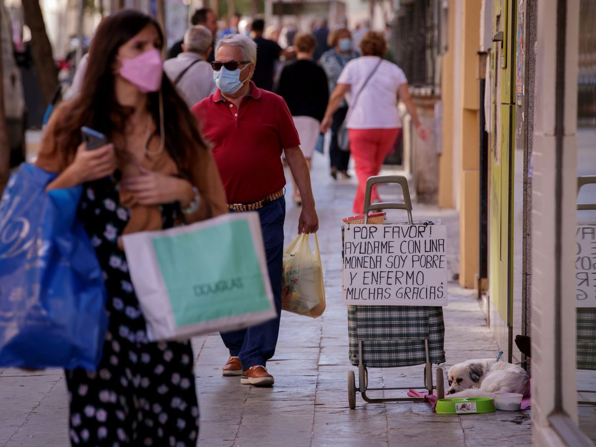 Foto: Una mujer pasea por el centro de Sevilla junto al cartel de una persona que solicita ayuda en la calle. (EFE/Julio Muñoz)
