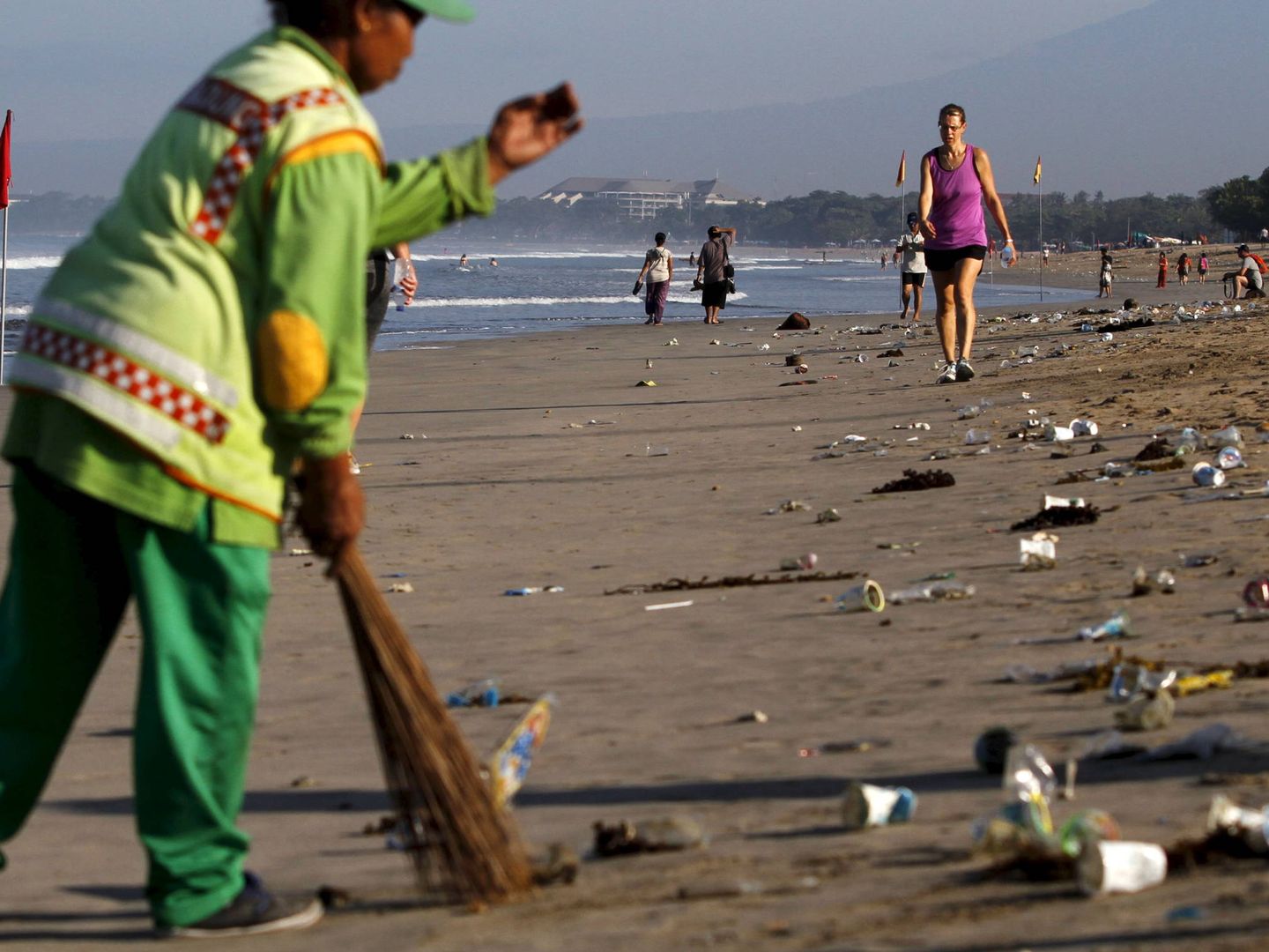 Turistas caminan junto a un trabajador que limpia la paya de Kuta, Bali. (Reuters)