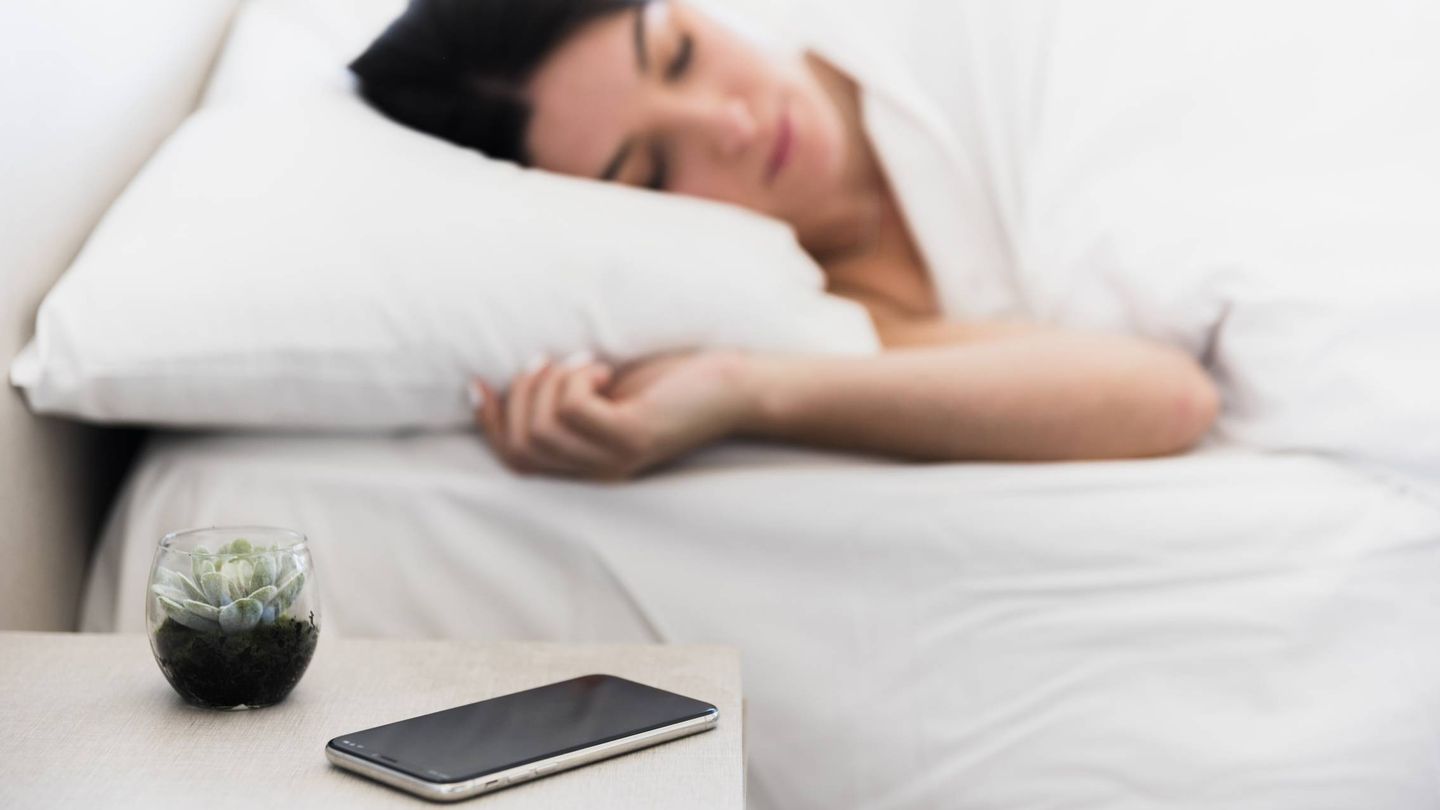 Una mujer duerme con su móvil y una planta en la mesilla de noche. (Freepik)