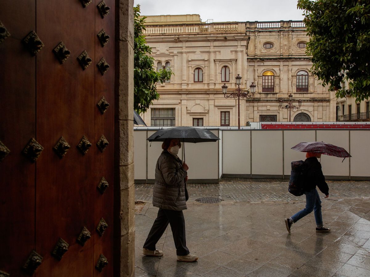 Foto: Dos mujeres con paraguas pasan junto a los palcos de Semana Santa instalados ante el Ayuntamiento de Sevilla. (EFE)