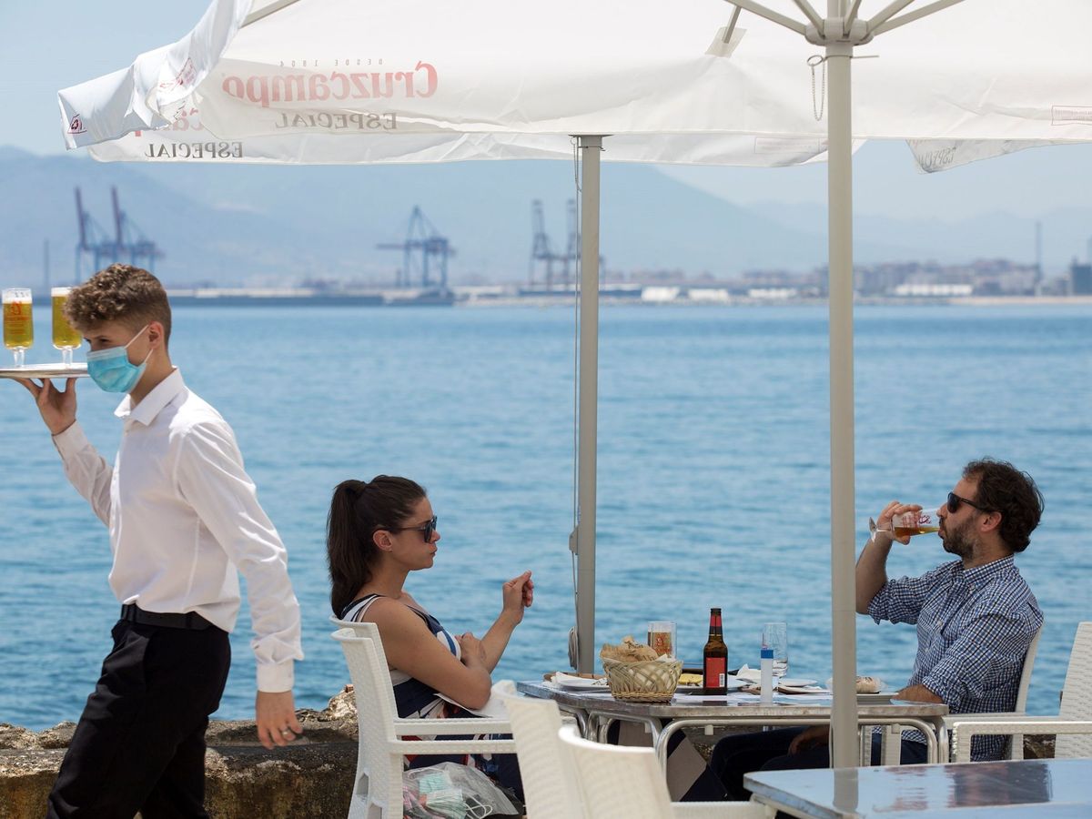 Foto: Un camarero con mascarilla pasa ante una pareja que almuerza en un restaurante de Málaga. (EFE)
